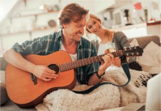 A senior man playing the guitar with a woman sitting next to him on a couch