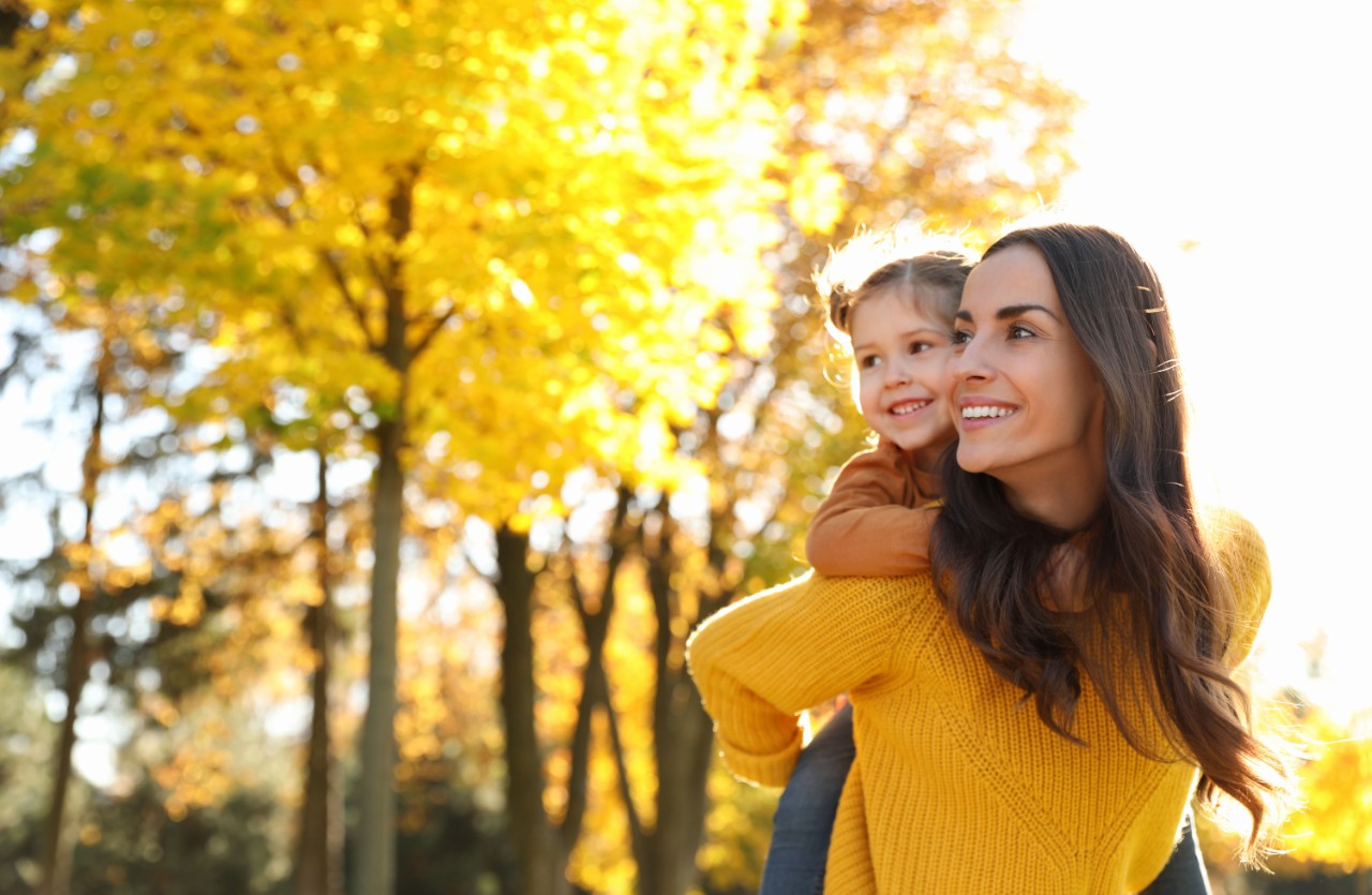 Mother carrying her little girl on her shoulders against an autumn backdrop