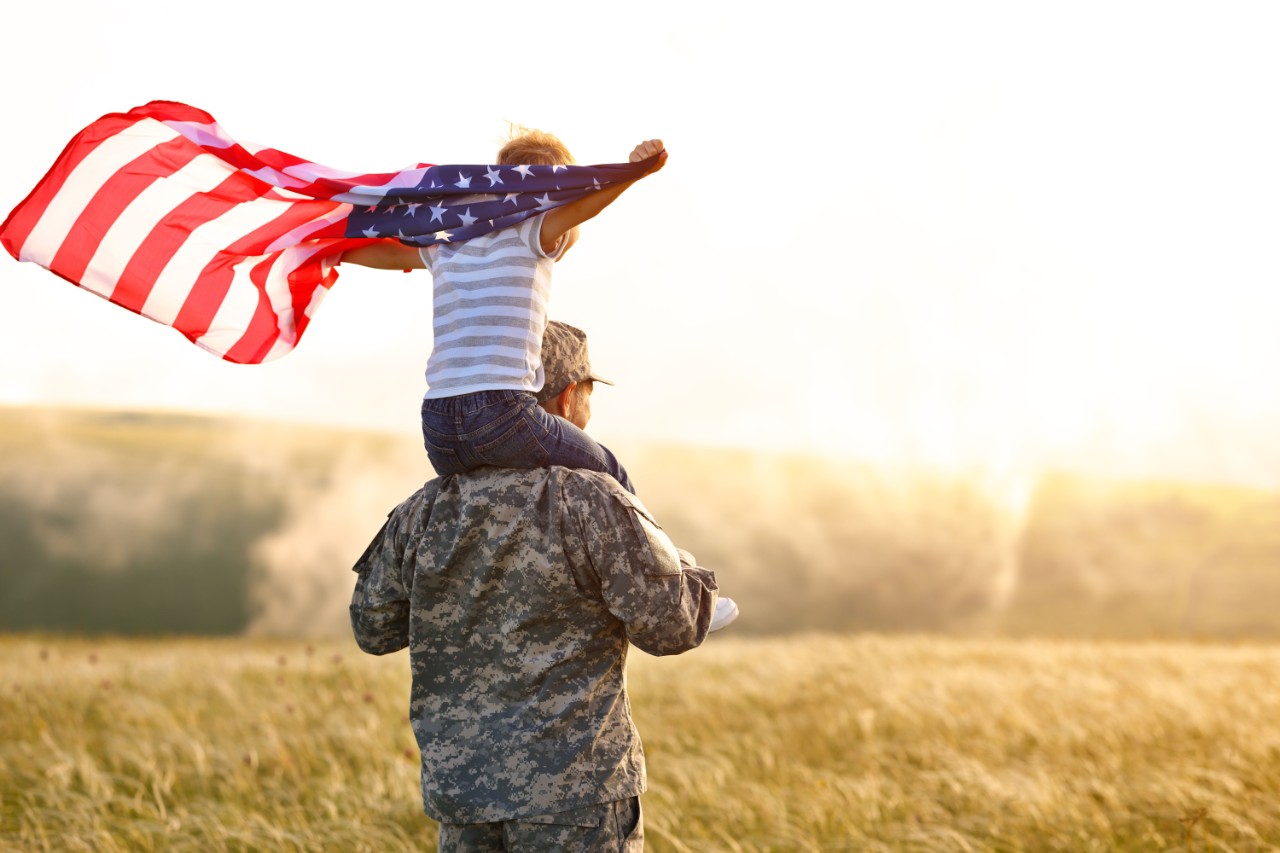 A veteran with his son on his shoulders waving the American flag