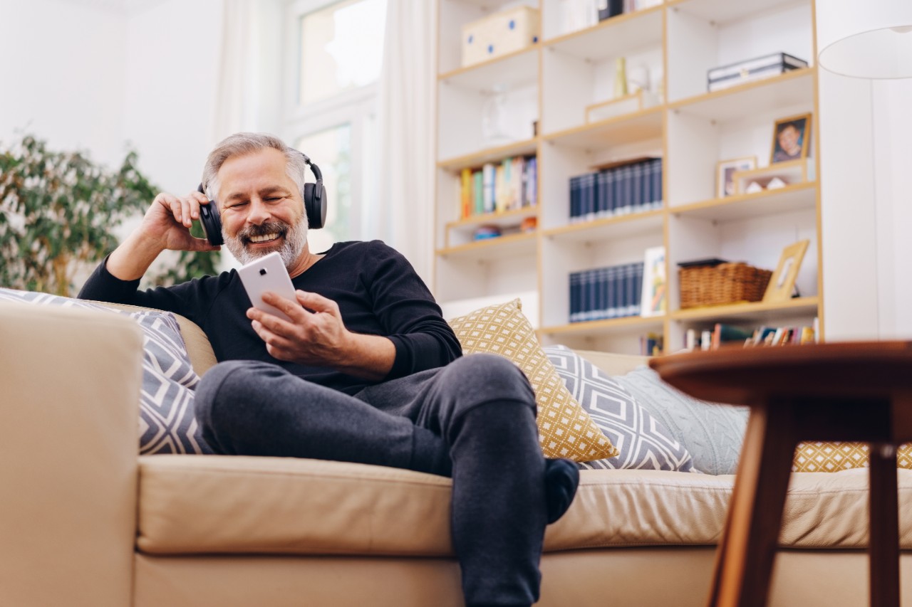 Grey hair gentleman on couch with headphones and phone smiling